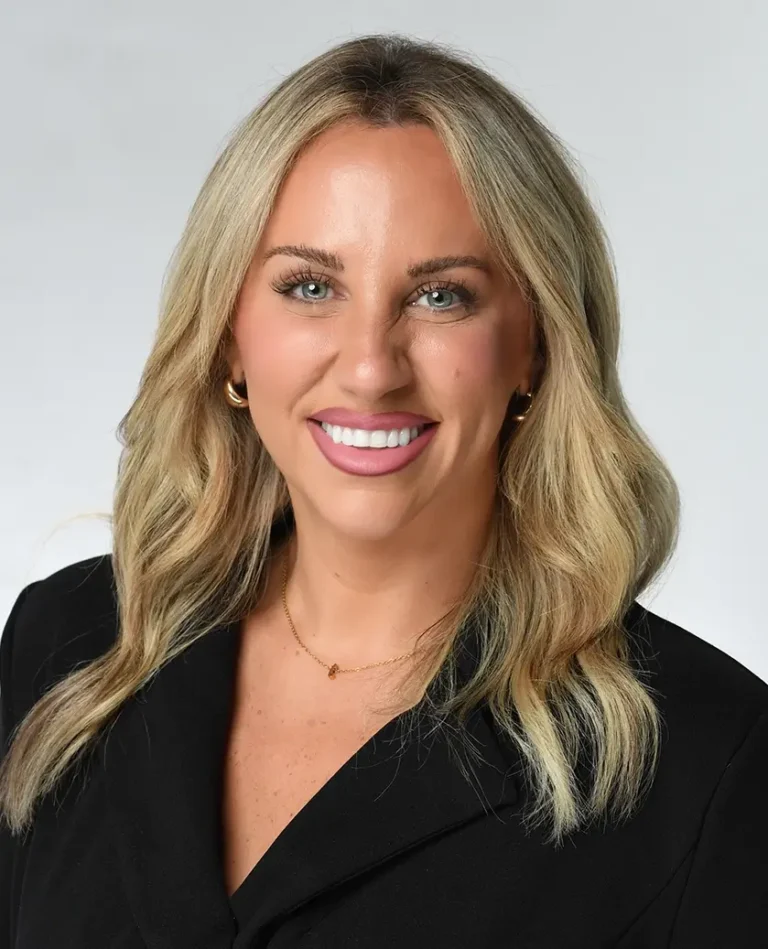 A woman with long blonde hair smiles at the camera. She is wearing a black top, gold hoop earrings, and a necklace, with a plain light background.