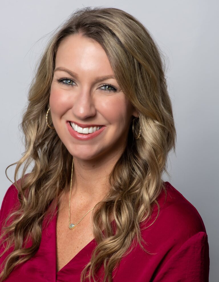 Woman with wavy blonde hair, wearing a red blouse, gold hoop earrings, and a gold necklace, smiling in front of a plain light gray background.
