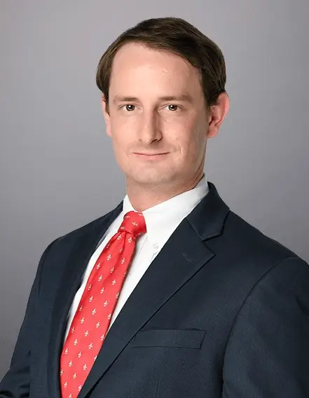 Man in a dark suit, white shirt, and red patterned tie poses for a professional headshot against a plain gray background.