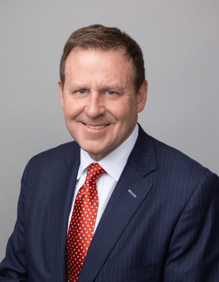 A man in a dark pinstripe suit, white shirt, and red polka dot tie smiles in front of a plain gray background.