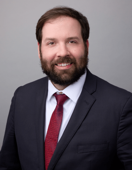 Bearded man in a dark suit, white shirt, and red tie poses against a plain gray background.