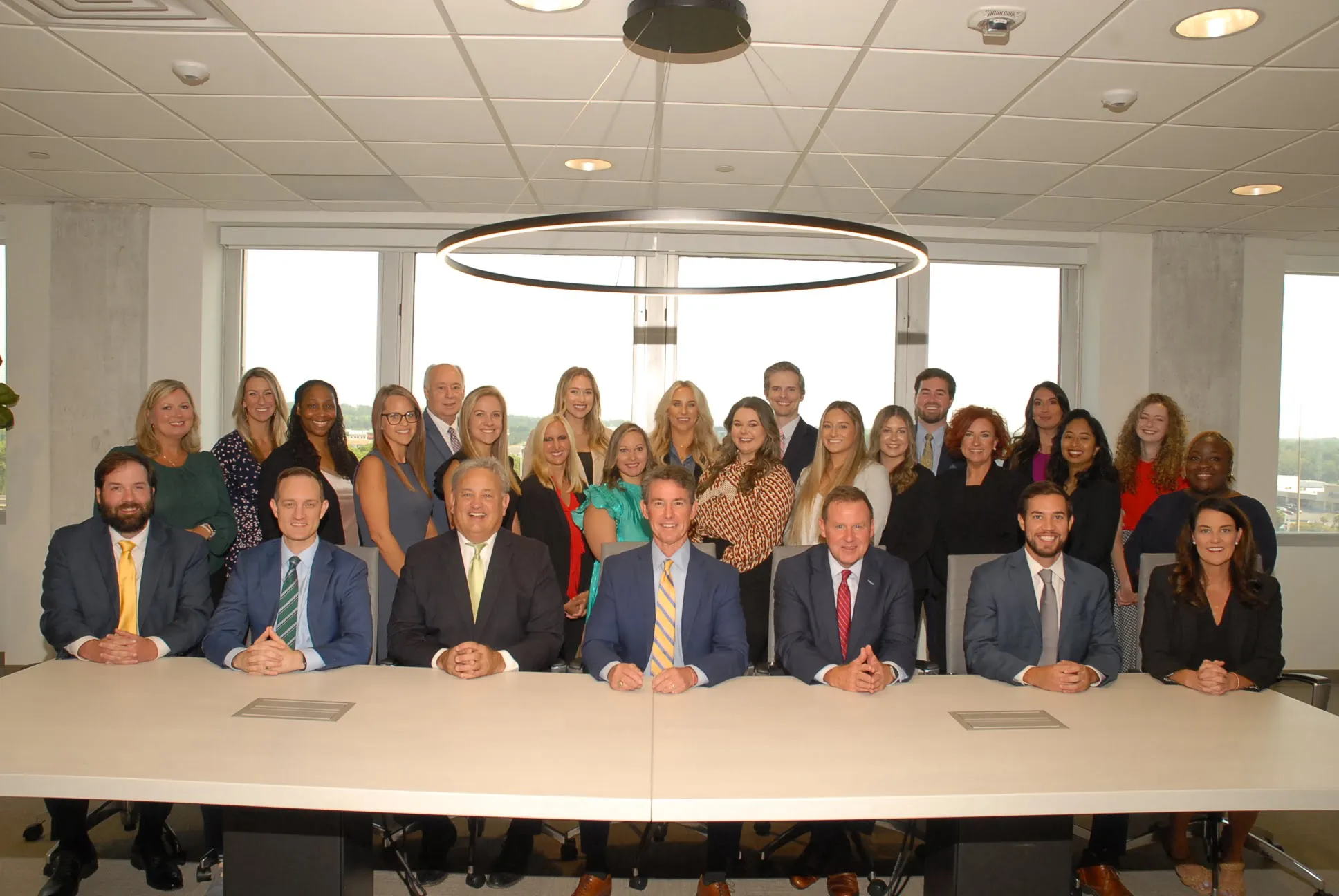 A group of twenty-three professionally dressed people pose for a group photo in a modern conference room with a large window.
