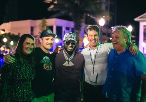 Five people stand together outdoors at night, smiling with arms around each other in a well-lit area with palm trees and buildings in the background.