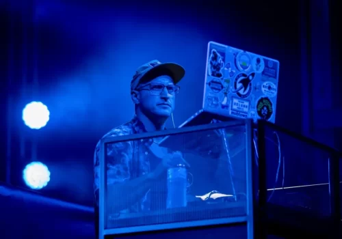 A person wearing a cap and glasses stands behind a laptop covered in stickers on a stage with blue lighting.