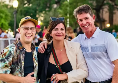 Three people stand outdoors smiling, with a crowd and trees in the background; two men flank a woman, and all appear casually dressed.