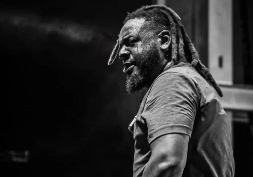 A man with dreadlocks and a beard, wearing a t-shirt, appears sweaty and intense, standing indoors. The image is in black and white.